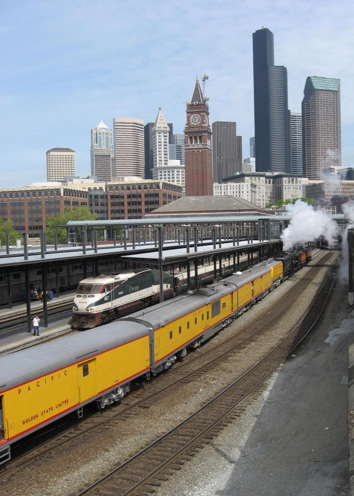 Panoramic view of yellow steam train in King Street station with Seattle skyline visible in background