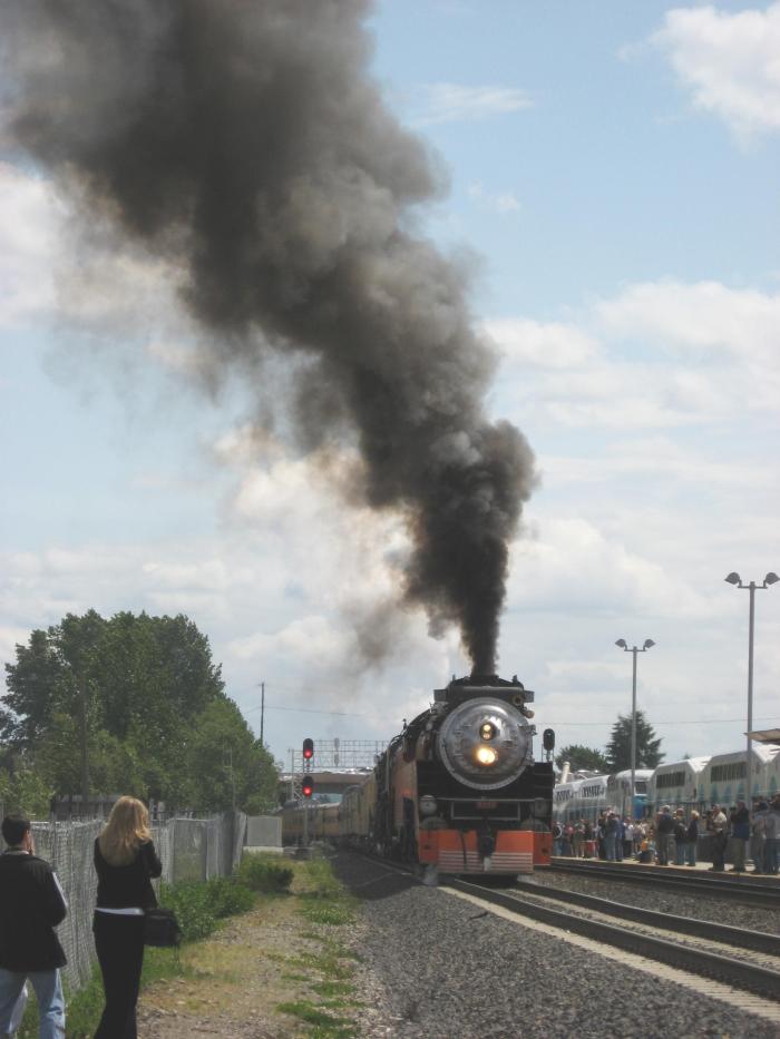 Steam locomotive approaching station with crowd on platform to right